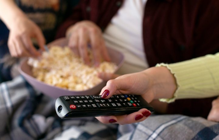 Hand som håller i en fjärrkontroll med händer i popcornskål i bakgrunden